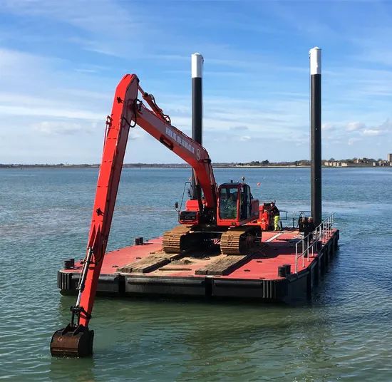 En röd grävmaskin på en pontonpråm utför arbete i skärgården under en klarblå himmel.